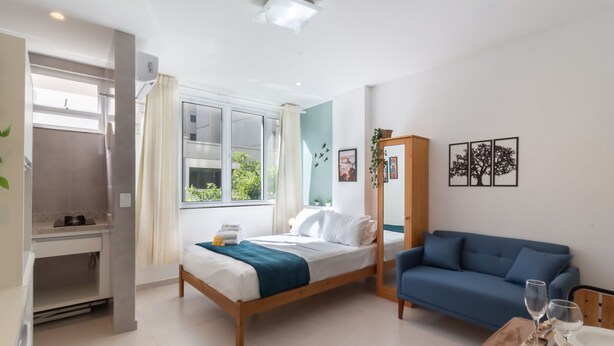 Ein modernes Schlafzimmer mit Bett, blauem Sofa, Esstisch mit Tellern und Gläsern sowie einem Fenster mit Vorhängen.