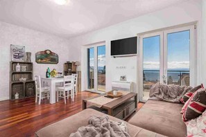 A living room with a wooden floor, a sofa, a dining table with chairs, a TV mounted on the wall, and large windows with a view of the sea. - 2 bedroom Apartment Victoria with panoramic views (Ičići)