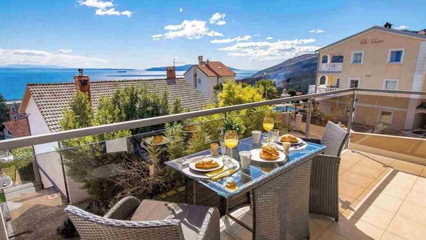 Ein Balkon mit Frühstückstisch, der auf eine Küstenlandschaft mit Häusern und blauem Himmel blickt.