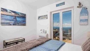Ein Schlafzimmer mit einem Bett, einem Gemälde, einem Koffer und Meerblick durch eine Schiebetür.