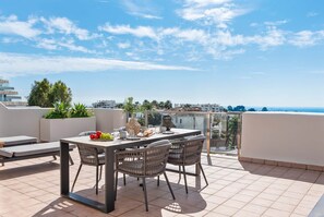 A terrace with a table and chairs, a view of the sea, and buildings in the background. - Luxe 2BR Penthouse | 55m² rooftop – Sea/Pool Views (Estepona)