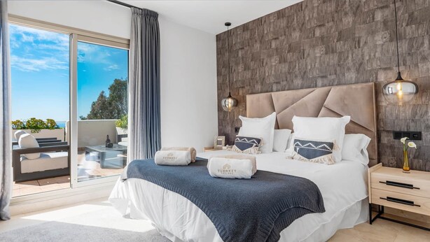 A modern bedroom with a large bed, a view of the outdoors, and a wooden accent wall. - Luxe 2BR Penthouse | 55m² rooftop – Sea/Pool Views (Estepona)