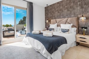 A modern bedroom with a large bed, a view of the outdoors, and a wooden accent wall. - Luxe 2BR Penthouse | 55m² rooftop – Sea/Pool Views (Estepona)