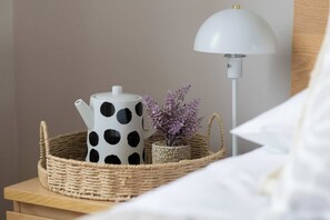Een nachtkastje met een witte lamp, een stipjespot, een geweven mand en een kleine potplant.