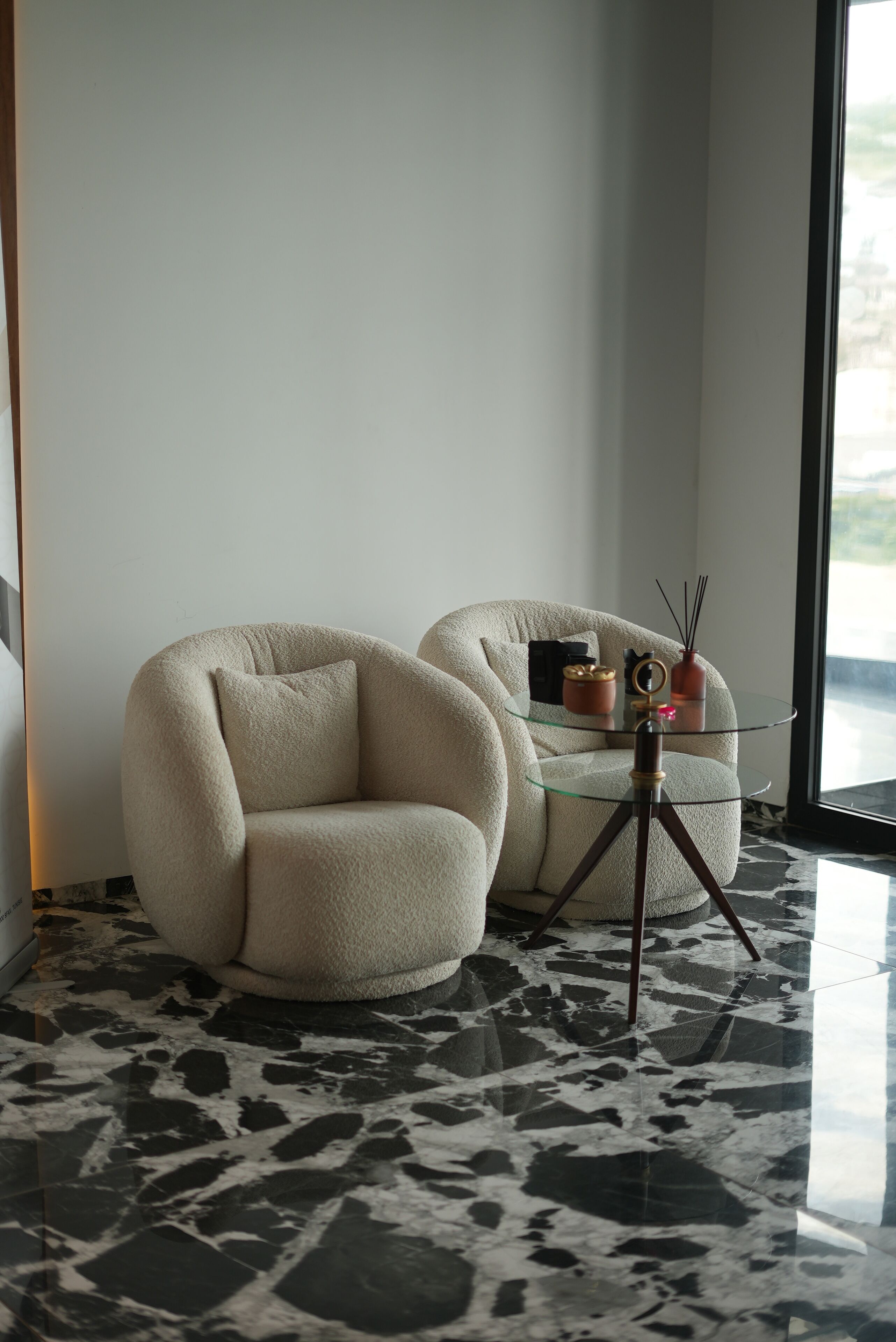 A modern room with a marble floor, two cream-colored armchairs, a glass coffee table, and a tripod lamp.
