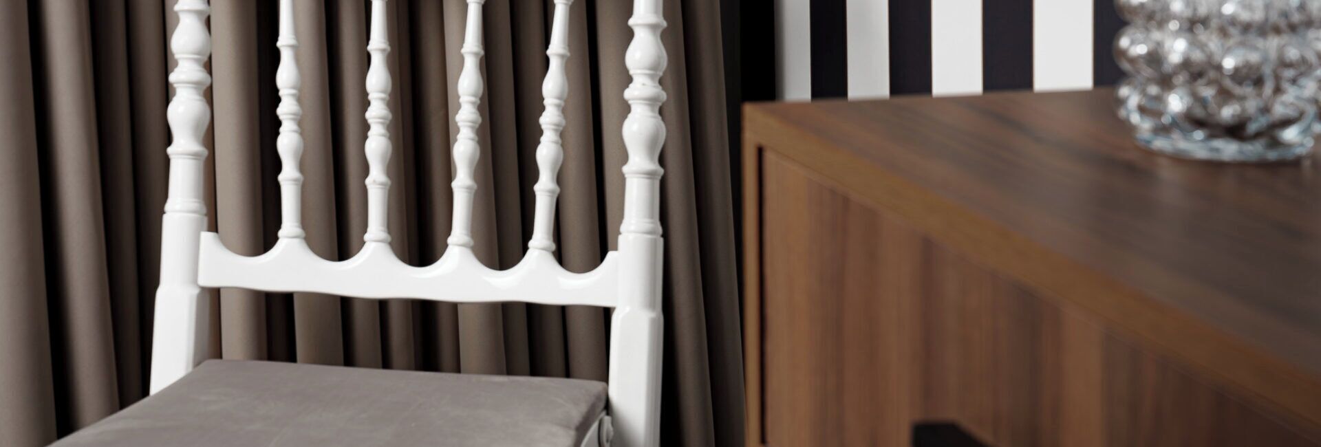 Una silla blanca con respaldo de madera y cojín gris, ubicada frente a una cortina oscura y una mesa de madera con un objeto de vidrio decorativo encima.