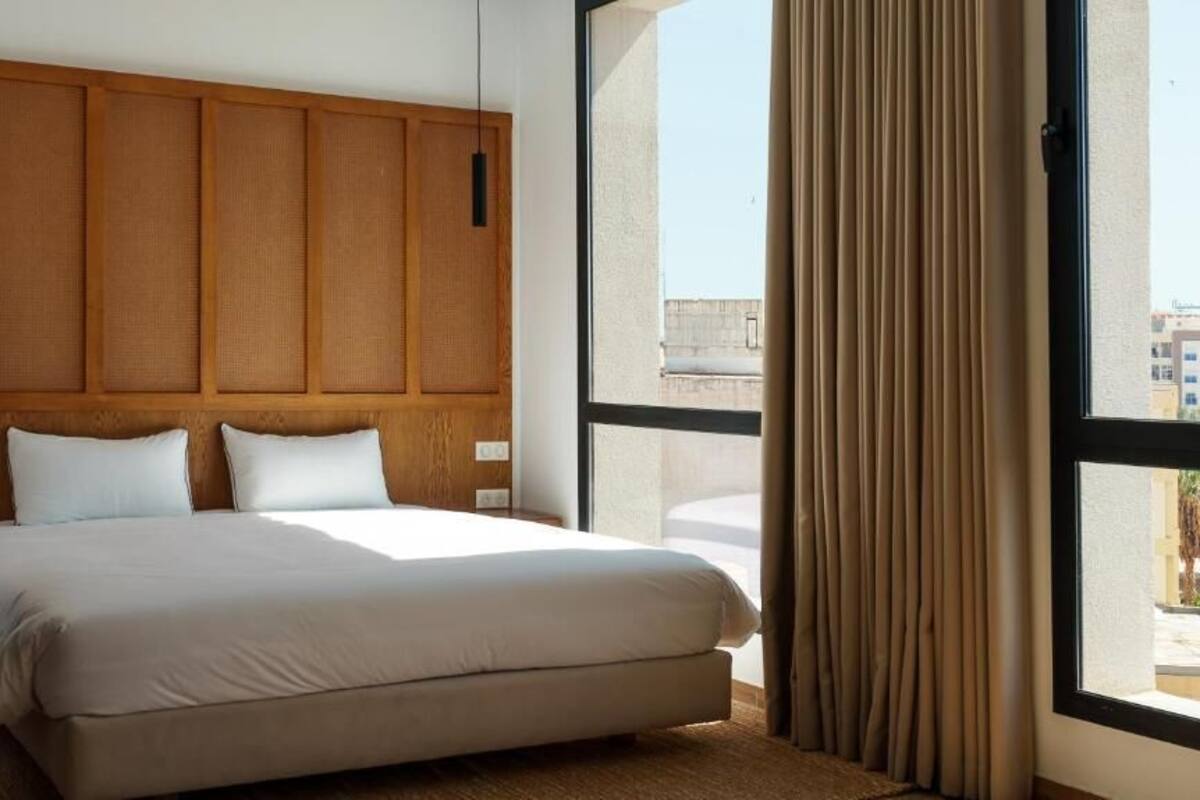 Habitación de hotel con una cama grande, cabecero de madera y vista a edificios a través del ventanal.