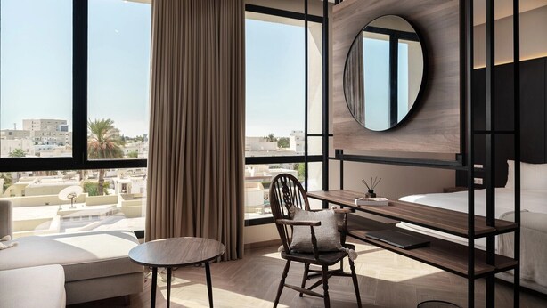 Una habitación de hotel moderna con un ventanal grande, un espejo redondo, un escritorio, una silla y una cama.