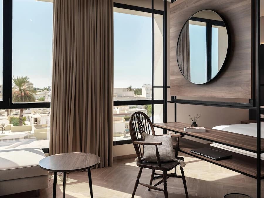 Una habitación de hotel moderna con un ventanal grande, un espejo redondo, un escritorio, una silla y una cama.