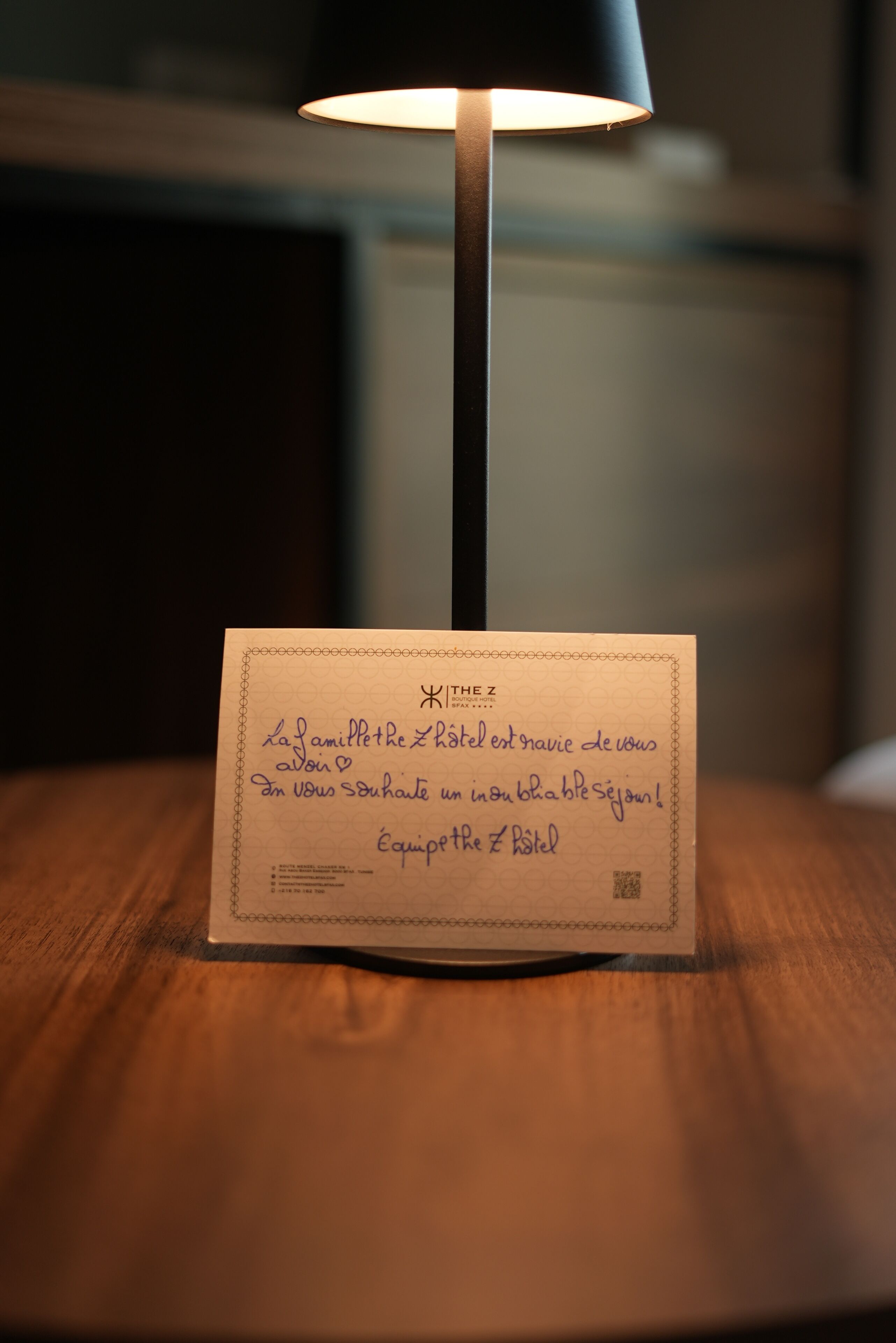 A handwritten note on a card placed on a wooden surface, illuminated by a lamp.