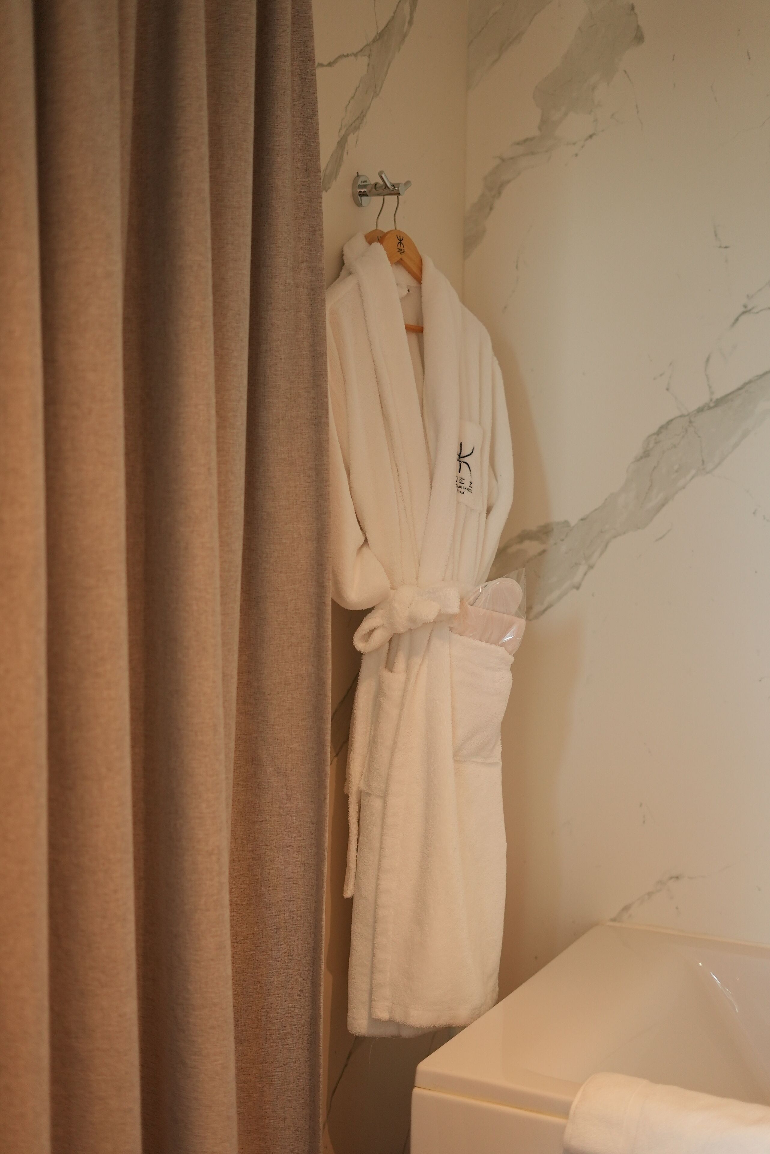 A bathroom with a marble wall, a white bathtub, and a white bathrobe hanging on a wooden hanger.