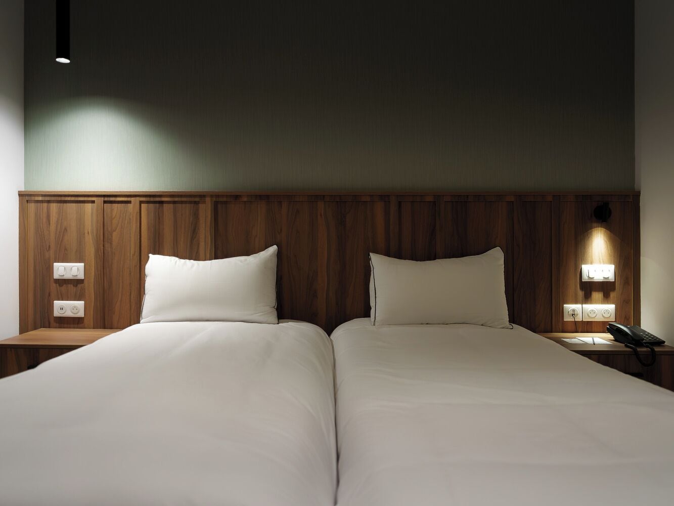 Cama doble con sábanas blancas, cabecera de madera, mesitas de noche con lámparas y un teléfono sobre la mesa derecha.