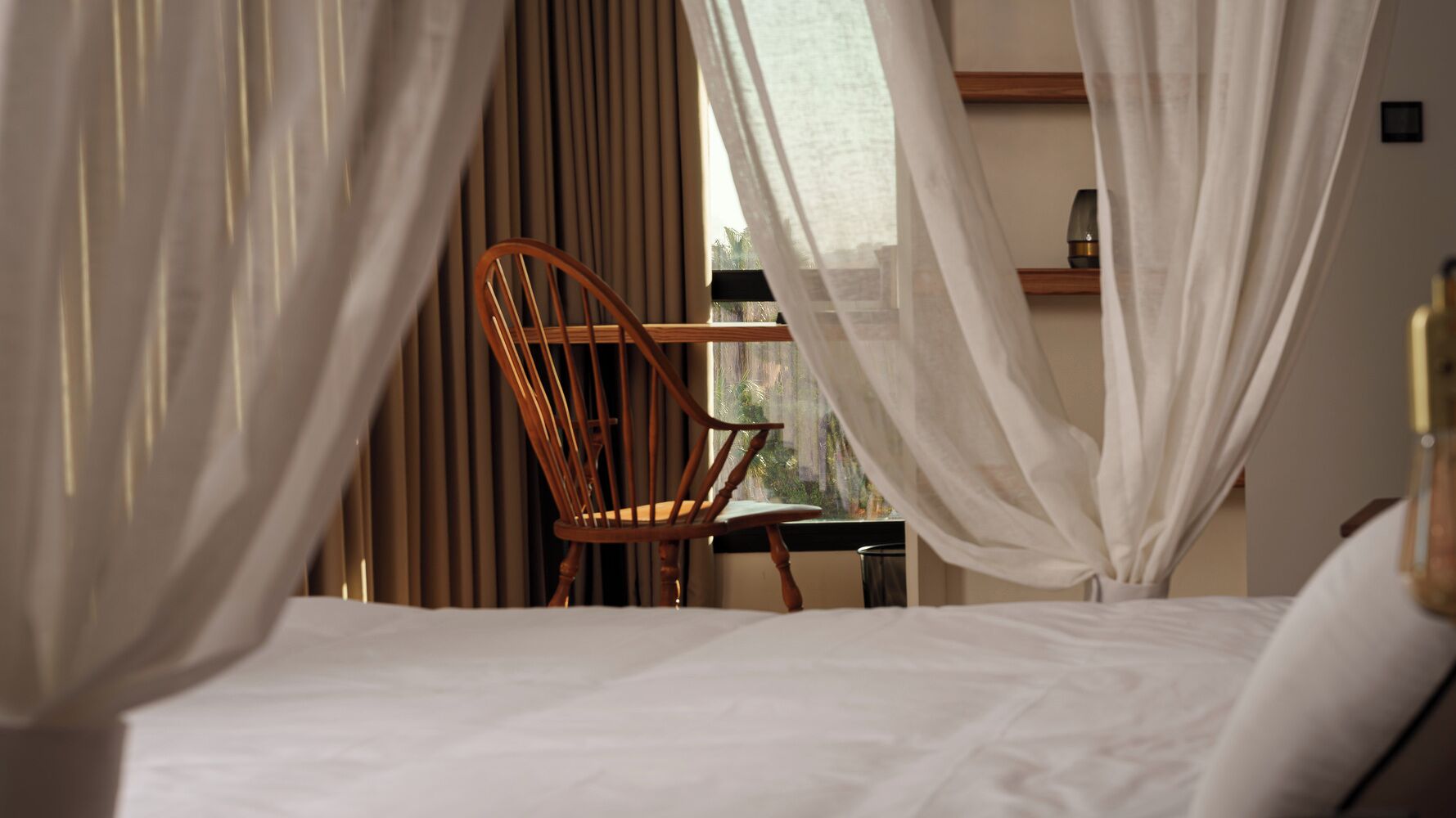 A hotel room with a bed, a wooden chair, a small table, and sheer curtains.