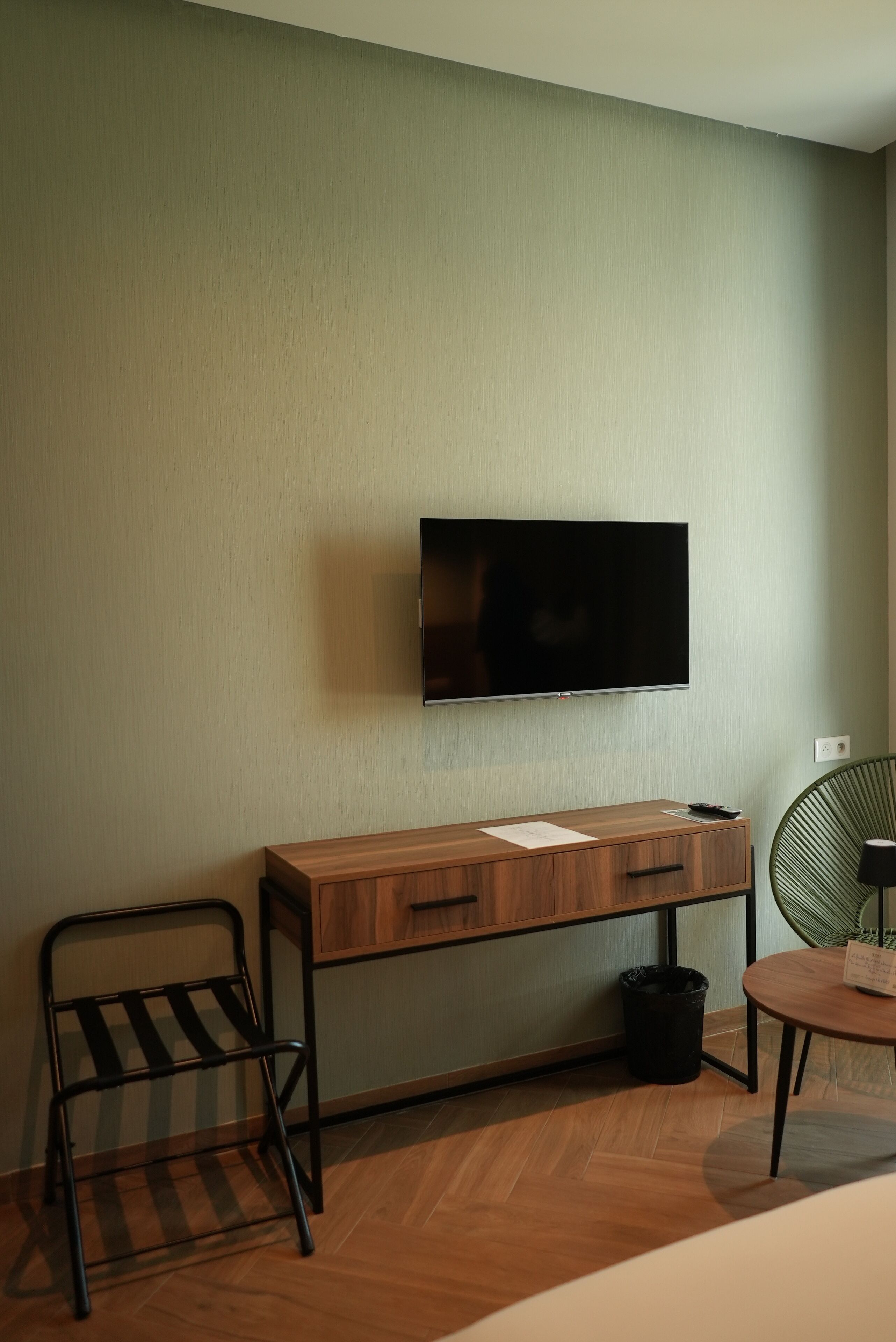 A hotel room with a wooden desk, a television mounted on the wall, a chair, a small table, and a trash bin.