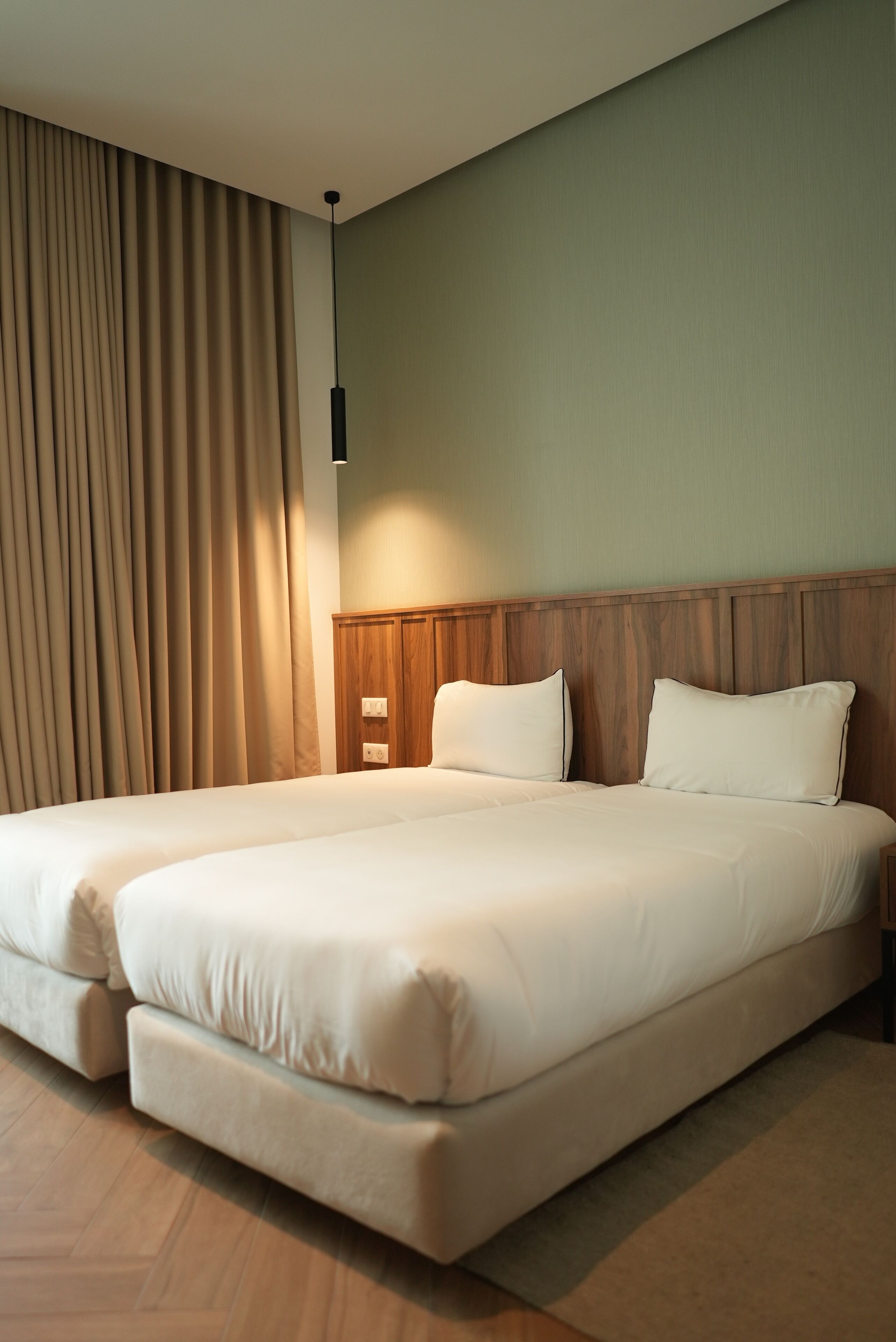 Una cama doble con cabecera de madera, cortinas color crema y un interruptor de luz en la pared.