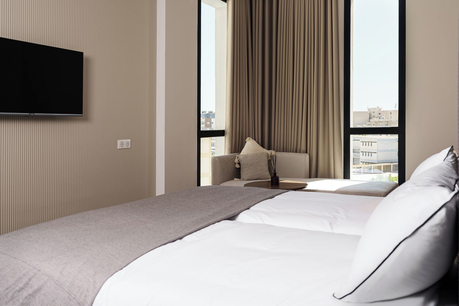 A hotel room with a bed, a TV mounted on the wall, and a view of buildings through large windows.