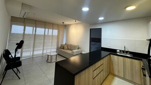 A modern kitchen with a black countertop, wooden cabinets, and a built-in oven. There is a seating area with a sofa and a small table, and a large window providing natural light. - Bright and Comfortable 2 Bedroom Apartment in Serena del Mar Cartagena (Cartagena)