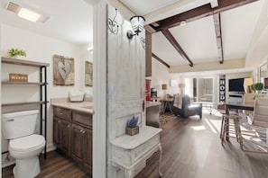 Een ruime woonkamer met hoge plafonds, zichtbare balken en een groot raam dat natuurlijk licht binnenlaat. Er is een keuken met moderne apparatuur en een eethoek met een bar.
