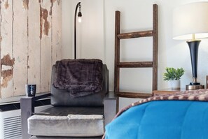 Ein Zimmer mit einem grauen Sessel, der mit einer dunklen Decke bedeckt ist, einer an der Wand stehenden Holzleiter, einer Stehlampe und einem kleinen Topfpflanzen auf einem Beistelltisch.