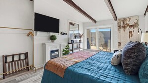 A bedroom with a bed, a TV mounted on the wall, a fireplace, and a view of the outdoors. - Driftwood Den – Condo for 2 (Kimberling City)