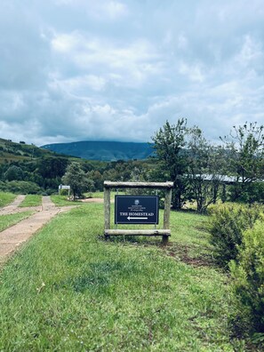 Schild für 'The Homestead' am GAMBATHI MOUNTAIN LODGE, das eine ländliche, naturnahe Umgebung mit einem Feldweg, der zum Lodge führt, kennzeichnet.