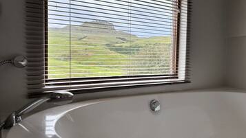 A bathroom with a large bathtub, a window with blinds, and a view of a green landscape.