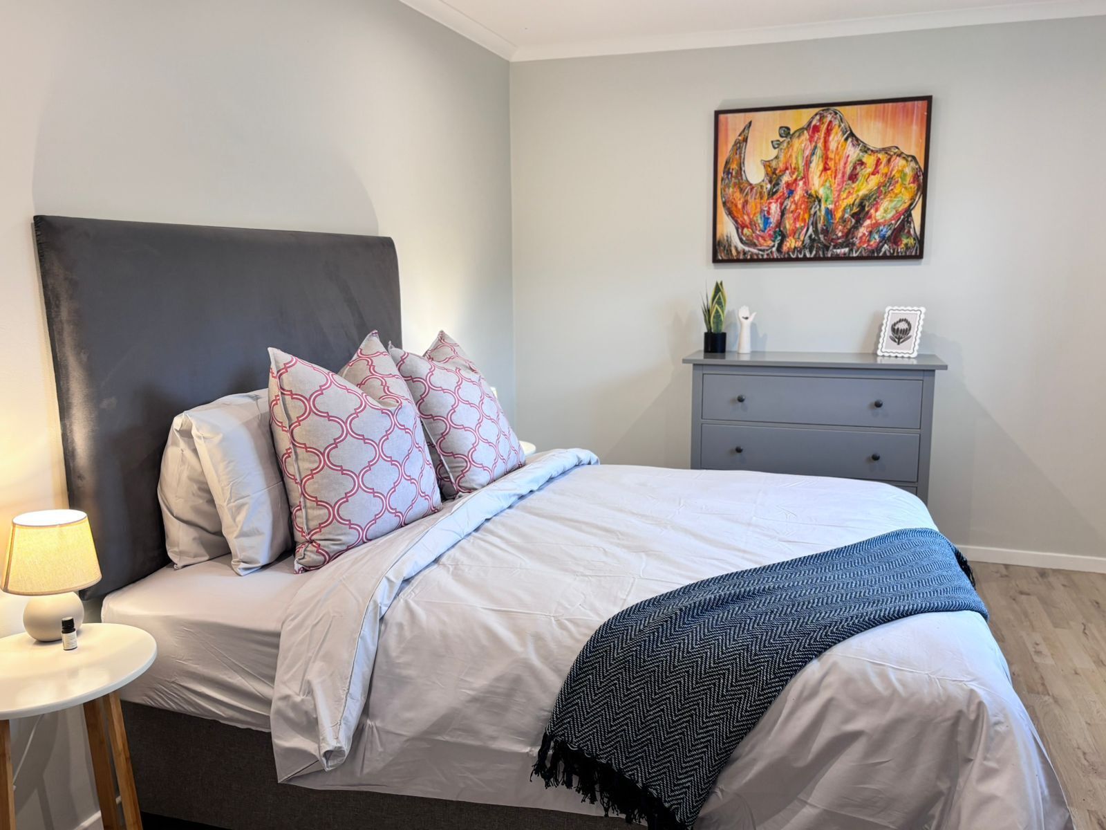 A neatly made bed with patterned pillows, a bedside table with a lamp, a gray dresser, and a colorful abstract painting on the wall.