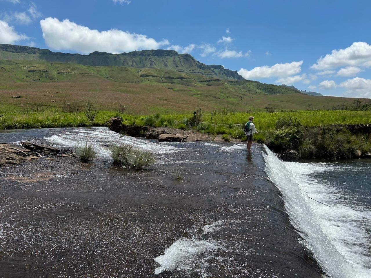 A person standing by a river with a mountainous backdrop.