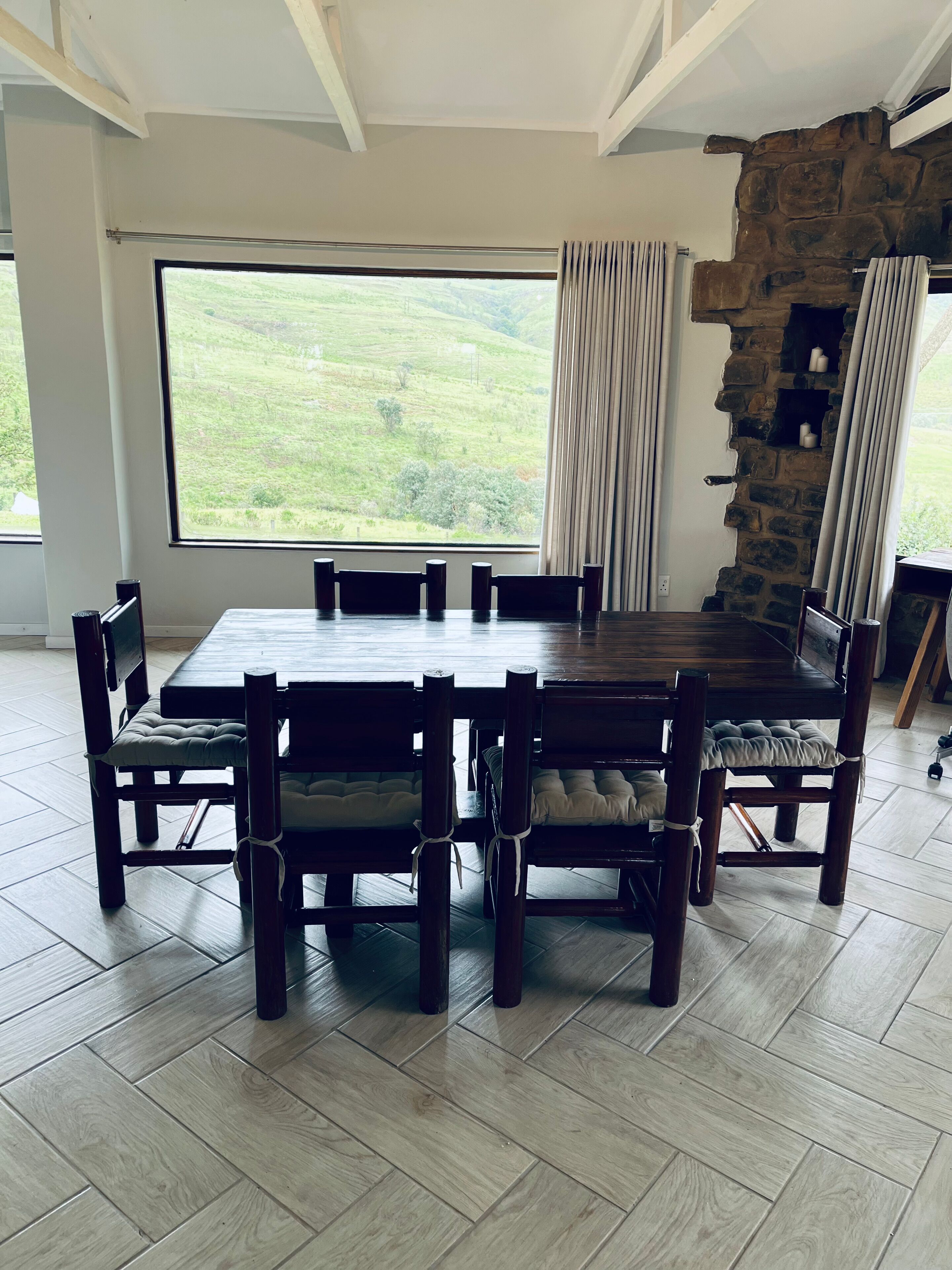 A dining area with a large table and chairs, a stone wall, and a view of a green landscape.