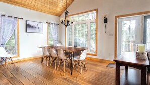 A dining area with a wooden table and chairs, large windows, and a chandelier.