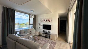 Una sala de estar moderna con un sofá, una mesa de centro y un televisor de pantalla plana. Hay un ventanal con cortinas, un cuadro en la pared y vistas al exterior.
