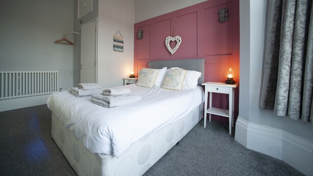 A neatly made bed with folded towels, a bedside table with a lamp, a heart-shaped decoration on the wall, and a radiator. - The Southport Two-Bed @ Stay Victoria Suites (Merseyside)