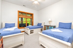 A room with three single beds, a wooden wardrobe, a desk with a lamp, and a window with a view of greenery.