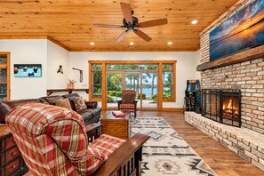 A spacious living room with a fireplace, a ceiling fan, and a large window offering a view of the outdoors.