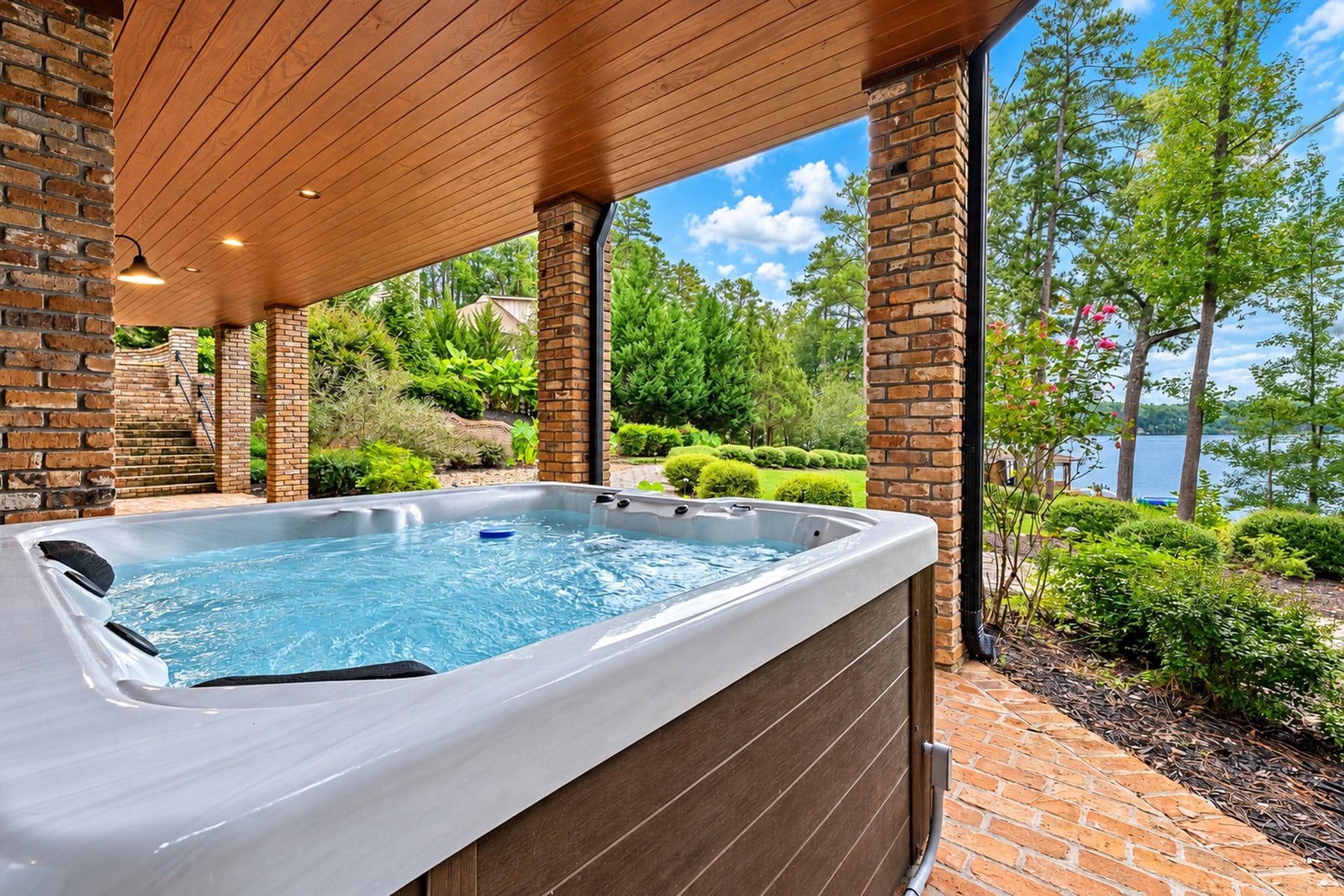 A hot tub under a wooden pergola with a brick wall, surrounded by lush greenery and a view of a lake.