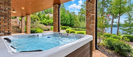 A hot tub under a wooden pergola with a brick wall, surrounded by lush greenery and a view of a lake.