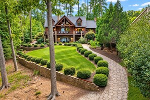 A two-story log cabin with a landscaped garden, a stone pathway, and a variety of trees and shrubs.