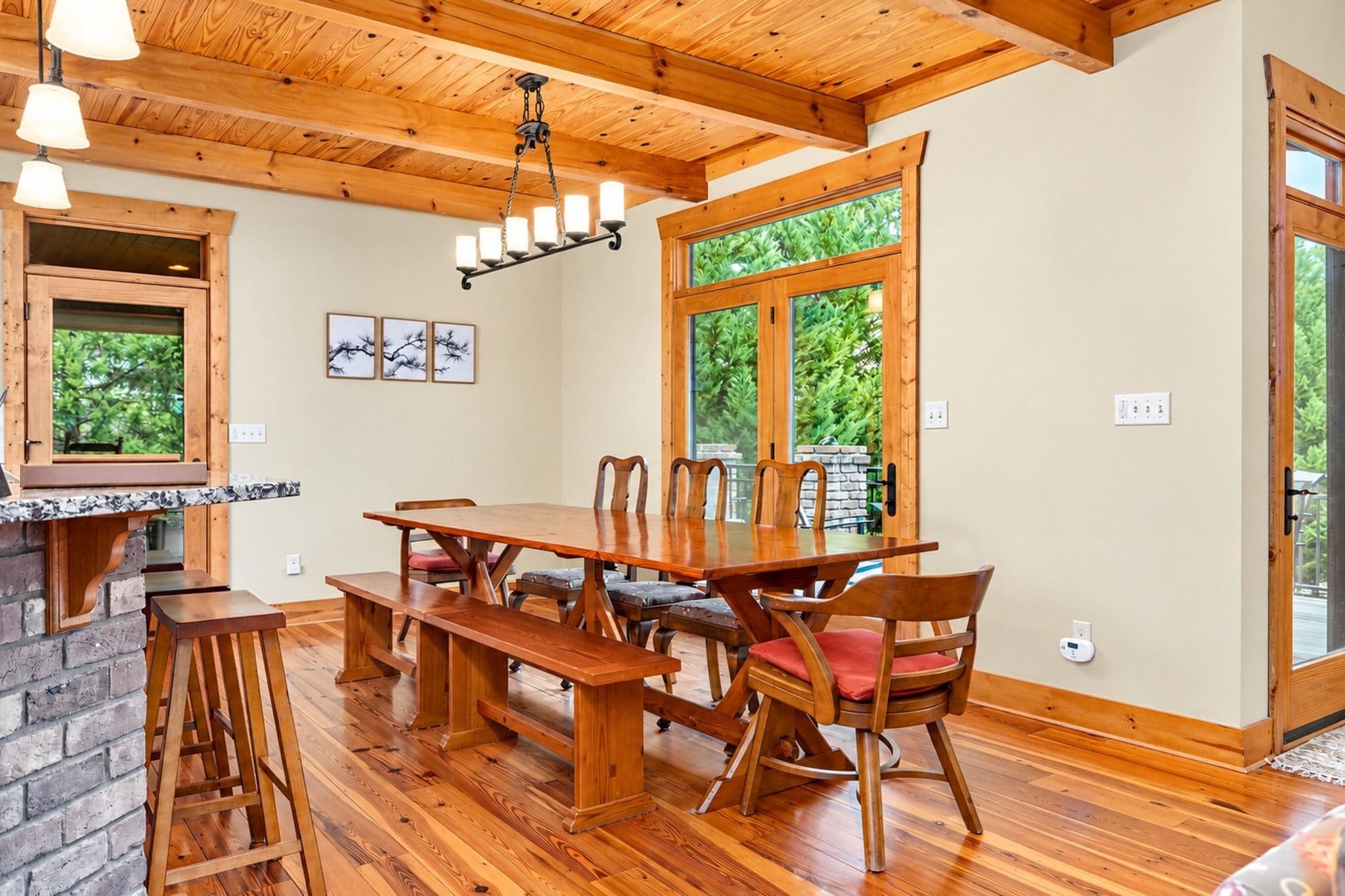 A dining area with a wooden table and chairs, a hanging light fixture, and a window with a view of greenery.