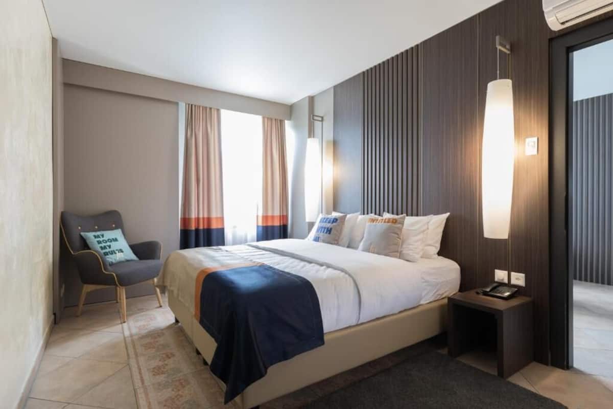 A modern hotel room with a large bed, a chair, a bedside table, and a wall-mounted lamp.