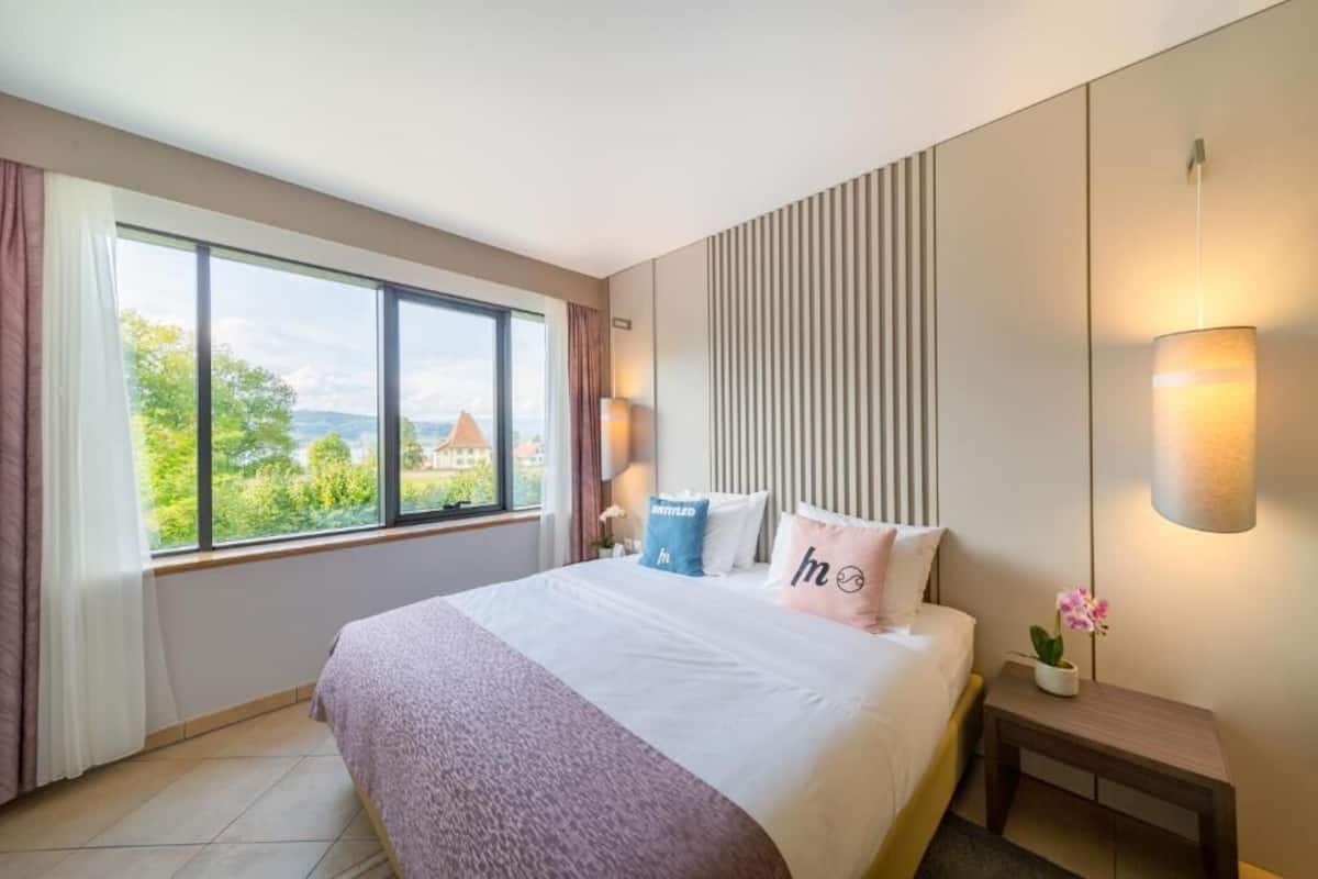 A modern hotel room with a large bed, a bedside table, a nightstand, a window with curtains, and a view of greenery and buildings outside.