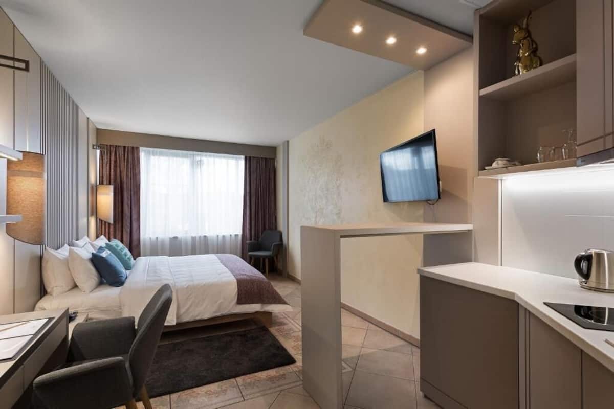 A modern hotel room with a bed, desk, and kitchenette.