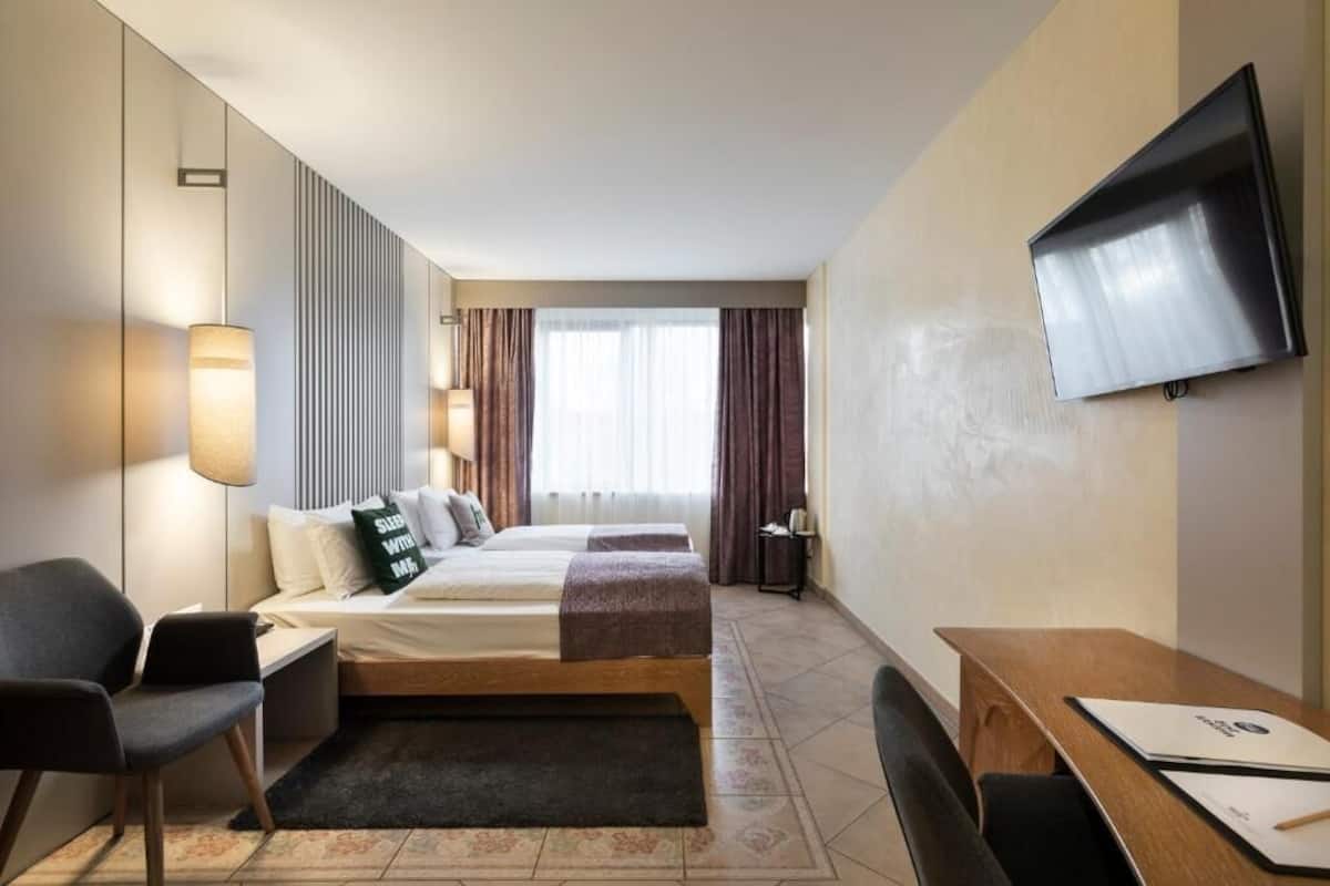 A hotel room with a bed, a desk, a chair, a TV, and a window with curtains.