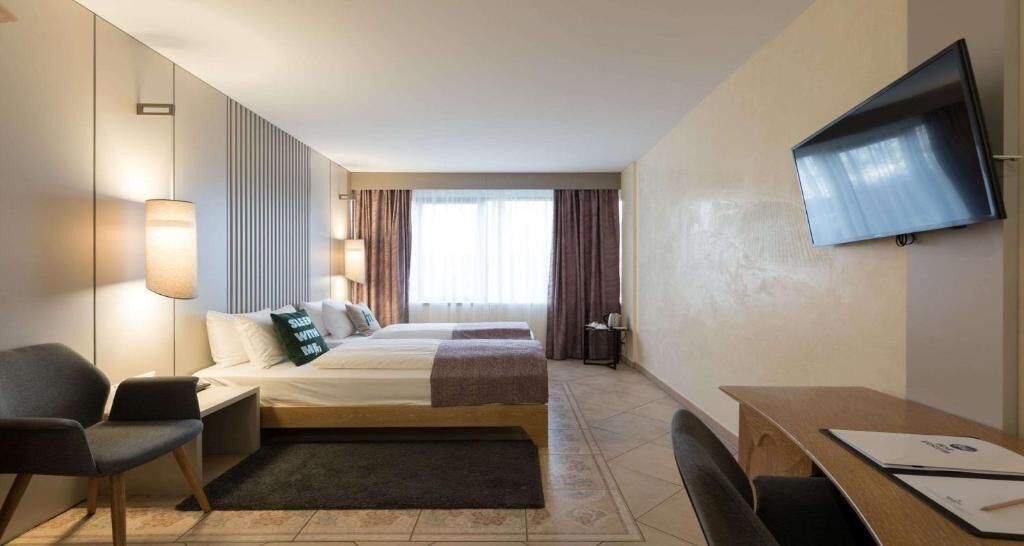Habitación de hotel con cama, dos mesitas de noche, un escritorio, una silla, televisor montado en la pared y una ventana con cortinas.