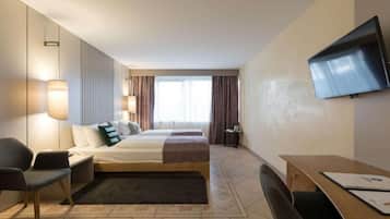 Habitación de hotel con cama, dos mesitas de noche, un escritorio, una silla, televisor montado en la pared y una ventana con cortinas.
