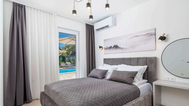 Ein modernes Schlafzimmer mit einem großen Bett, einem Nachttisch, einem Wandspiegel und Blick auf einen Außenpool-Bereich.