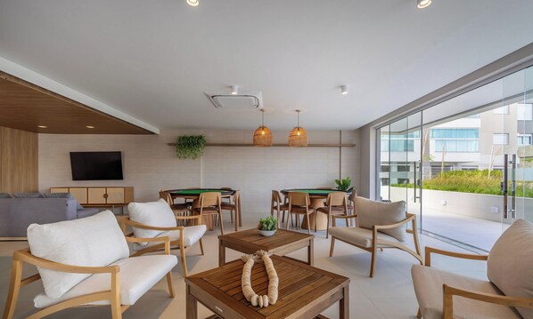 A modern lounge area with a pool table, comfortable seating, and a large window offering a view of the outside. - Cond. c/ Academia a apenas 200 m da Praia de Piçarras - PTCs (Picarras)