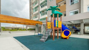 A playground with a slide and climbing structures, surrounded by a covered area and a building in the background. - Cond. c/ Academia a apenas 200 m da Praia de Piçarras - PTCs (Picarras)