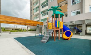 Un parque infantil con un tobogán y estructuras de escalada, rodeado de una zona cubierta y un edificio al fondo.