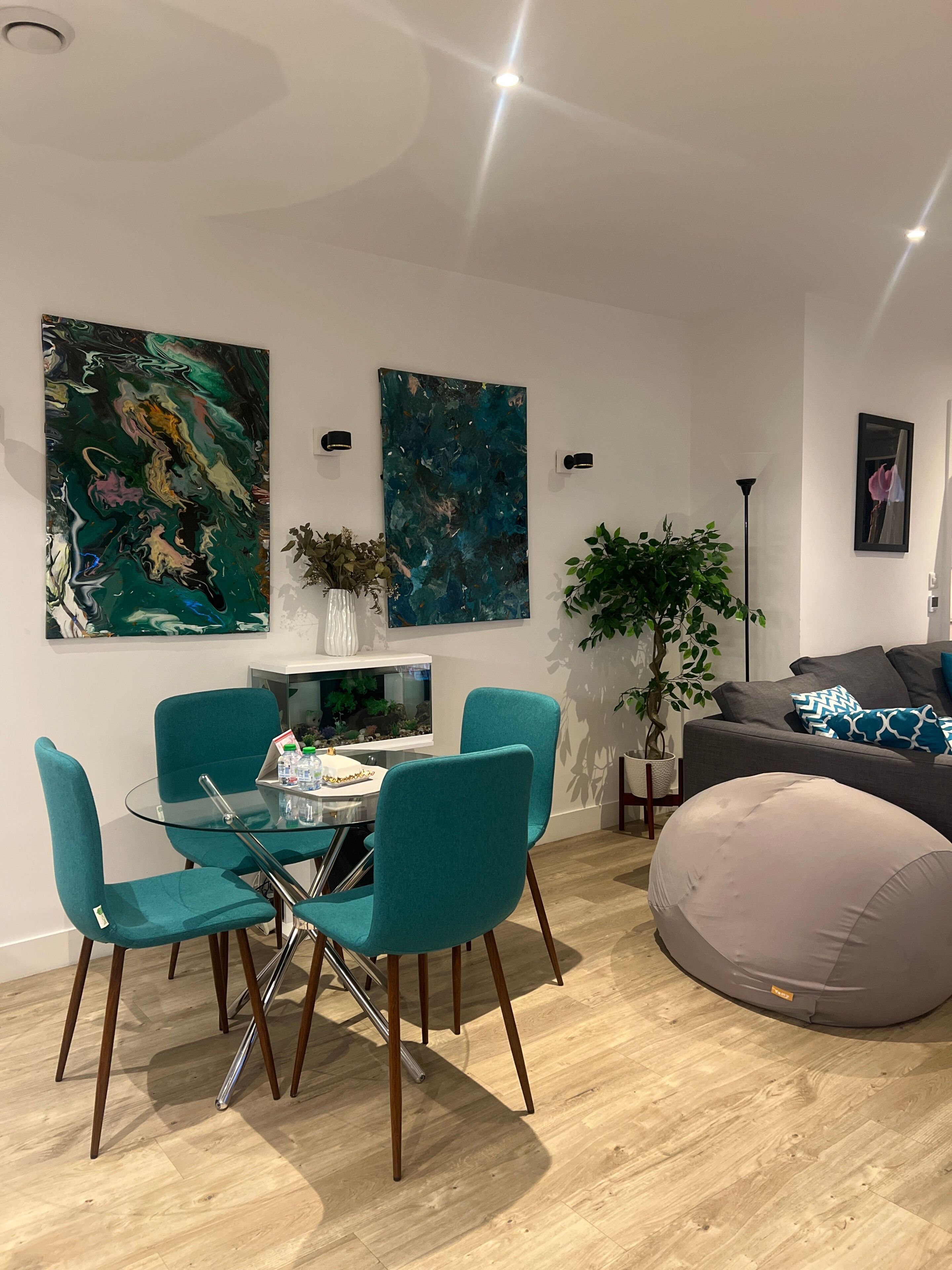 En moderne stue med teal farvede stole, et glas spisebord og en grå sofa.