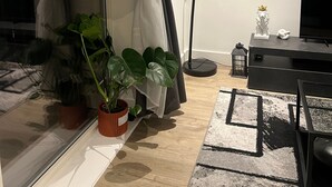 Un salon moderne doté d’une grande fenêtre, de rideaux légers, d’un lampadaire, d’un meuble TV noir et de plusieurs plantes en pot.